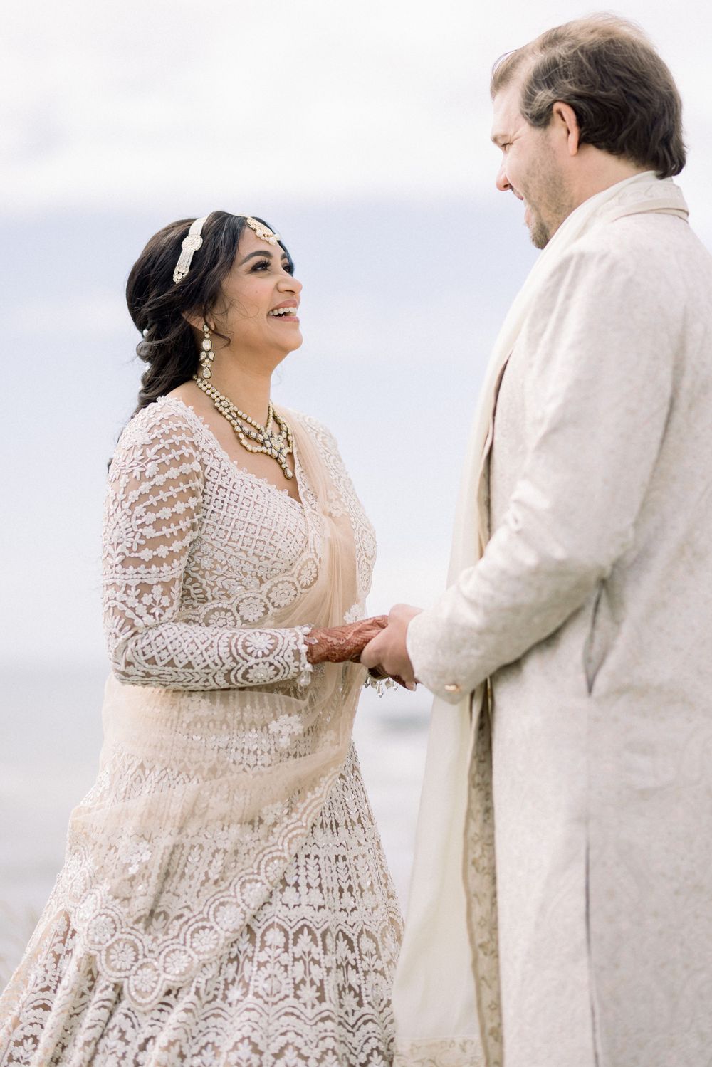 Couple in elegant cream-colored wedding attire sharing an intimate moment by the ocean.