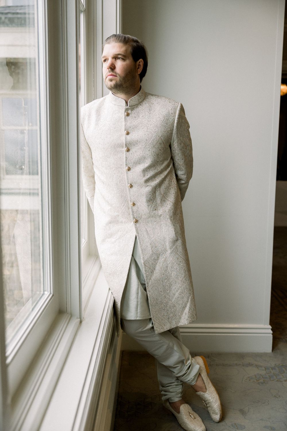 Groom in cream formal wear poses thoughtfully by bright windows.