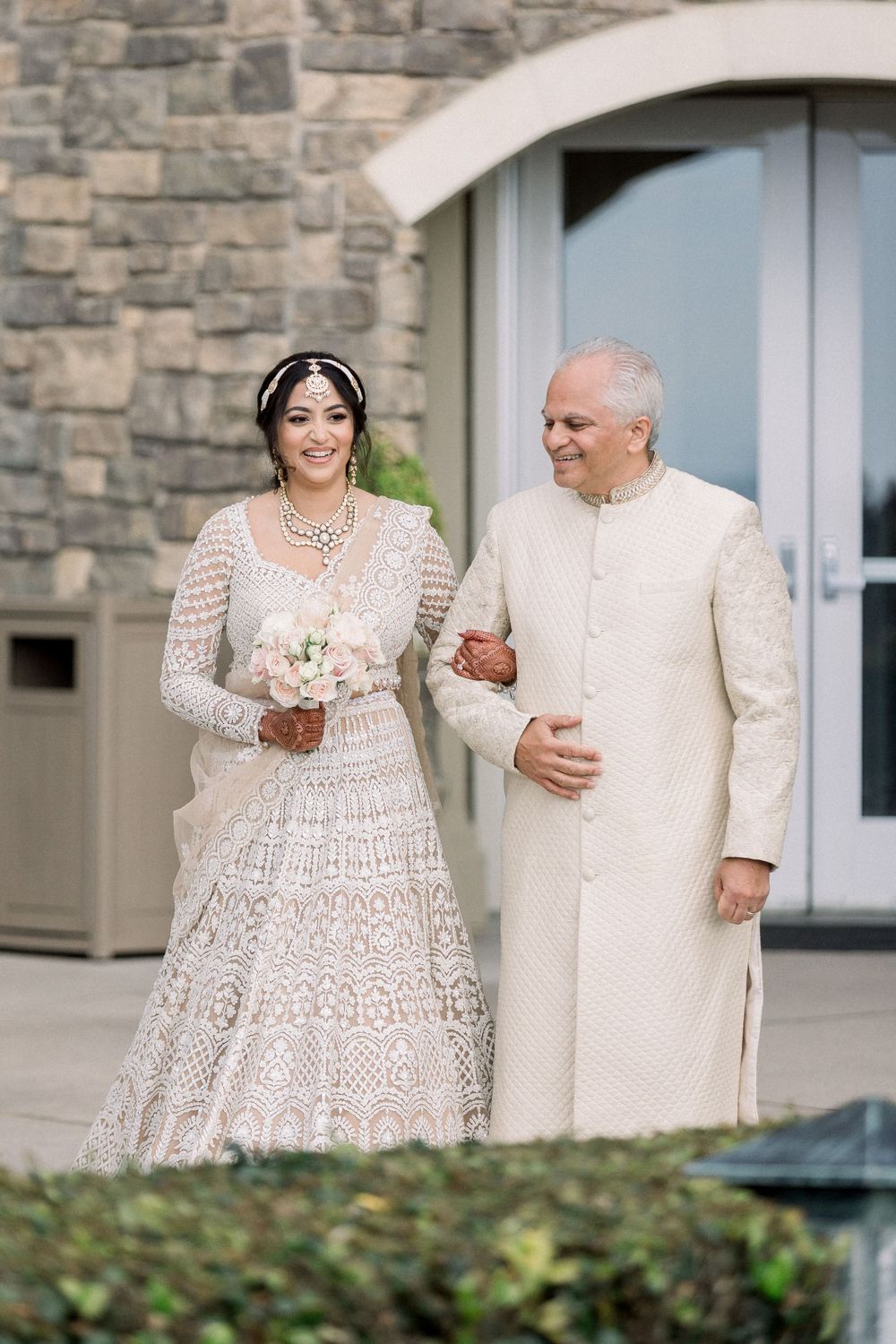 Wedding couple in cream and white outfits standing by stone building entrance.