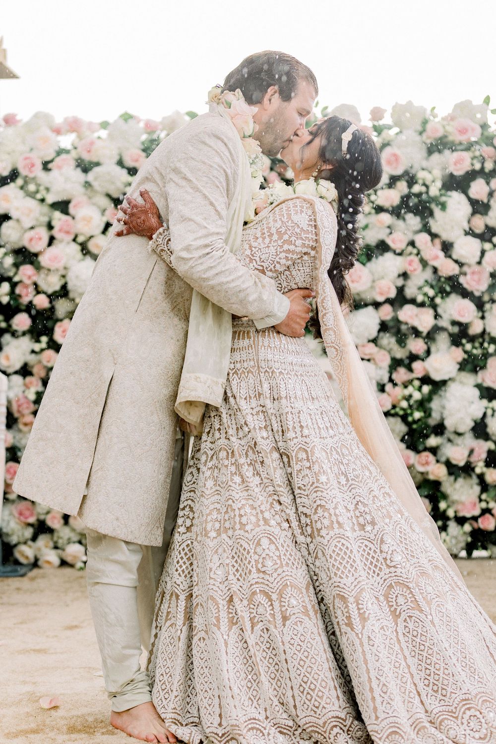 Couple in intricately detailed ivory wedding attire standing against floral wall.