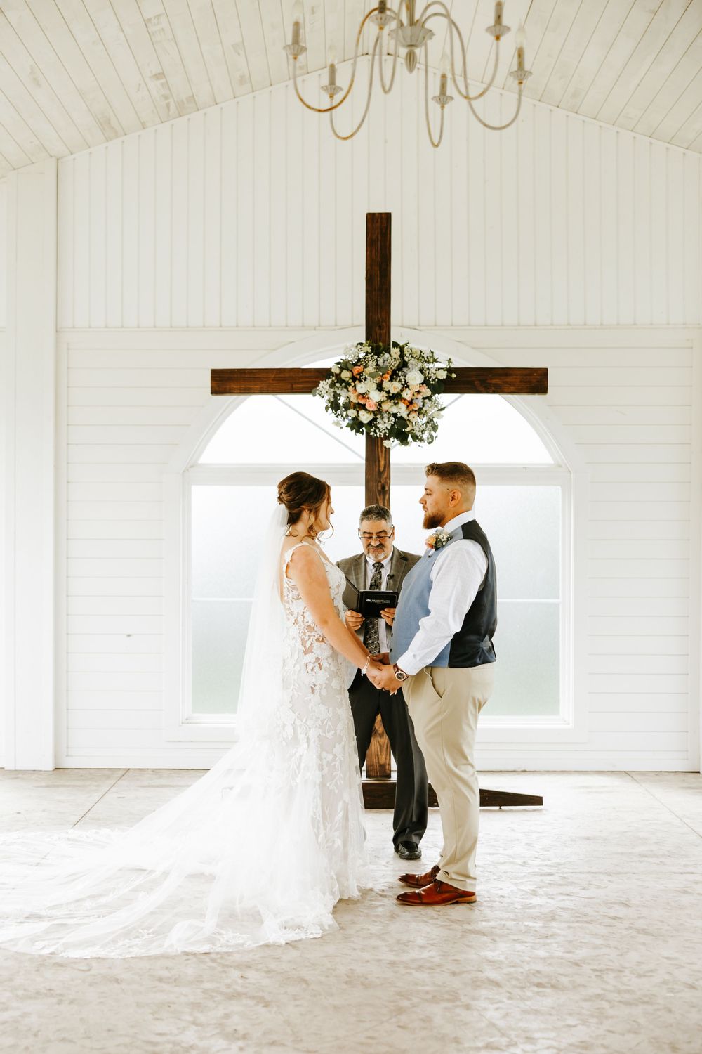 A wedding ceremony taking place in a white chapel with wooden cross and floral arrangement.