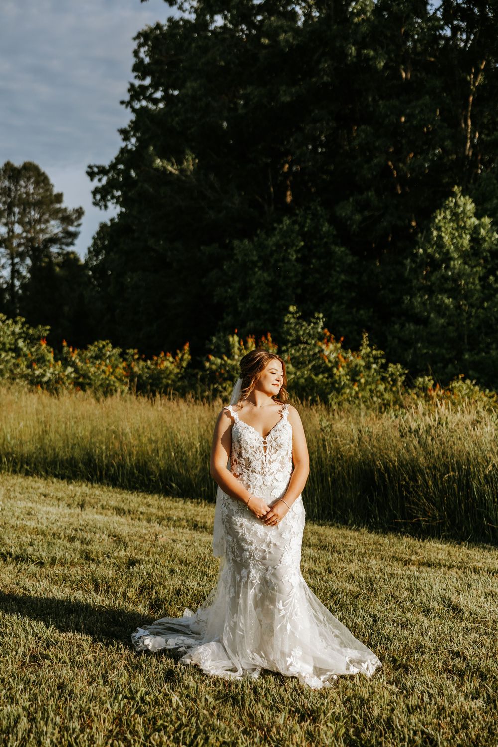 Person sitting in a white lace wedding dress on grassy hillside during golden hour with trees in background.