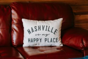 White decorative pillow with 'Nashville is my Happy Place' text on red leather couch.