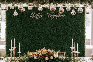 Green hedge wall wedding backdrop with 'Better Together' neon sign and floral arrangements.