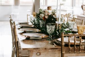 Elegantly set rustic wedding table with greenery garland centerpiece and gold chairs at an indoor reception.