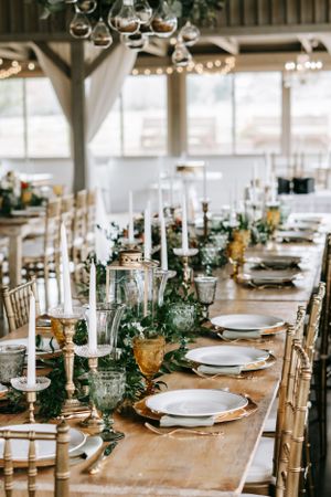 Romantic wedding reception table with white candles, gold accents and greenery runners on wooden tables.