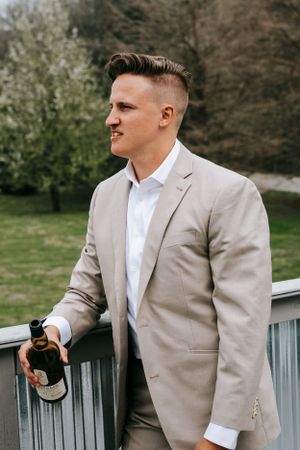A person in a beige suit stands on a balcony holding a drink.