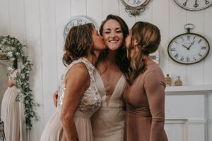 Friends in blush pink and white dresses share an intimate moment amid vintage clocks and floral decorations.