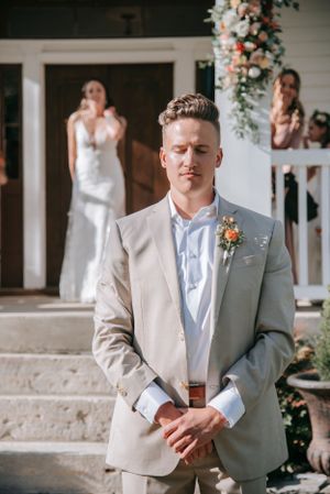 Groom in light gray suit stands confidently in front of wedding venue with floral arch.