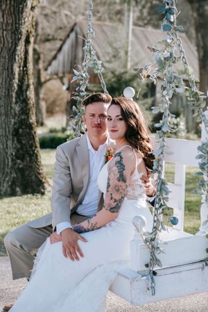 A romantic wedding photo on a white swing decorated with blue flowers and greenery in an outdoor garden setting.