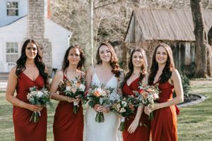 A bridal party in burgundy dresses holding floral bouquets poses in front of a rustic barn.