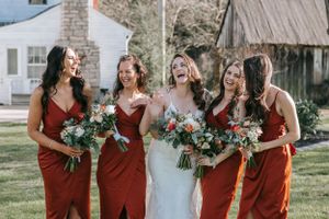 Bridesmaids in deep red dresses share a joyful moment while holding lush flower bouquets outdoors.