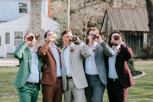 Groomsmen in colored suits laughing while taking drinks together outdoors.