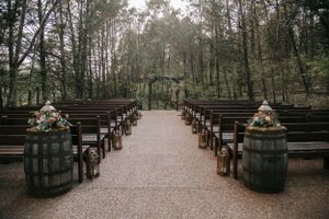 Rustic outdoor wedding ceremony setup with wooden barrels and benches in wooded setting.