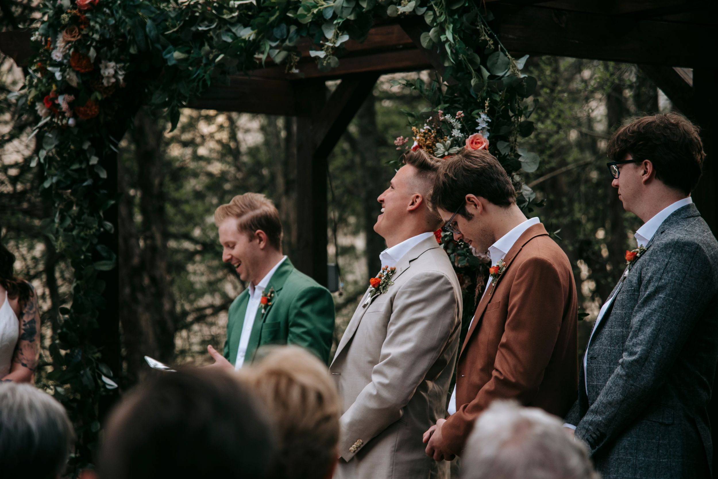 Wedding party stands under floral arch during outdoor forest ceremony in stylish suits of green, tan and brown.