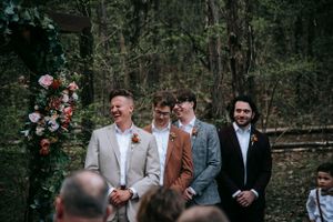 Groomsmen in varied colored suits stand together during outdoor forest wedding ceremony.