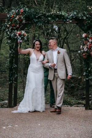 Joyful newlyweds celebrate under floral arch during outdoor forest wedding.