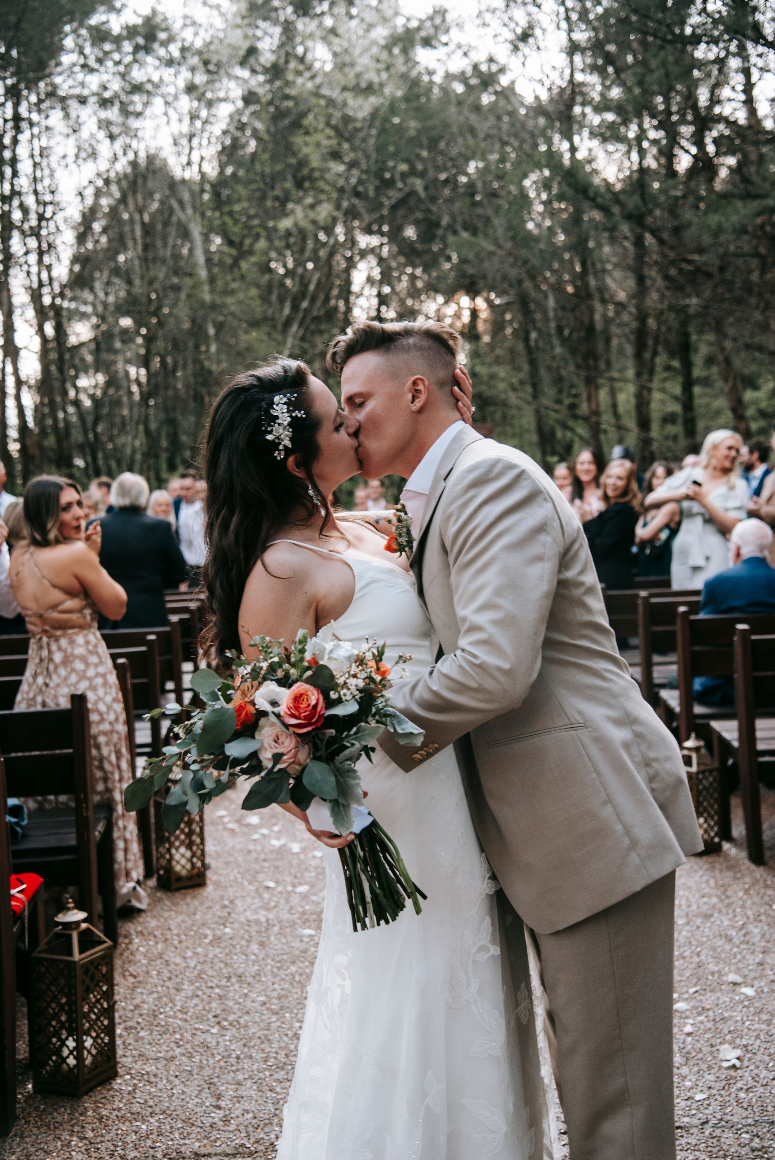 Newlyweds share romantic kiss while holding bouquet during outdoor woodland ceremony.