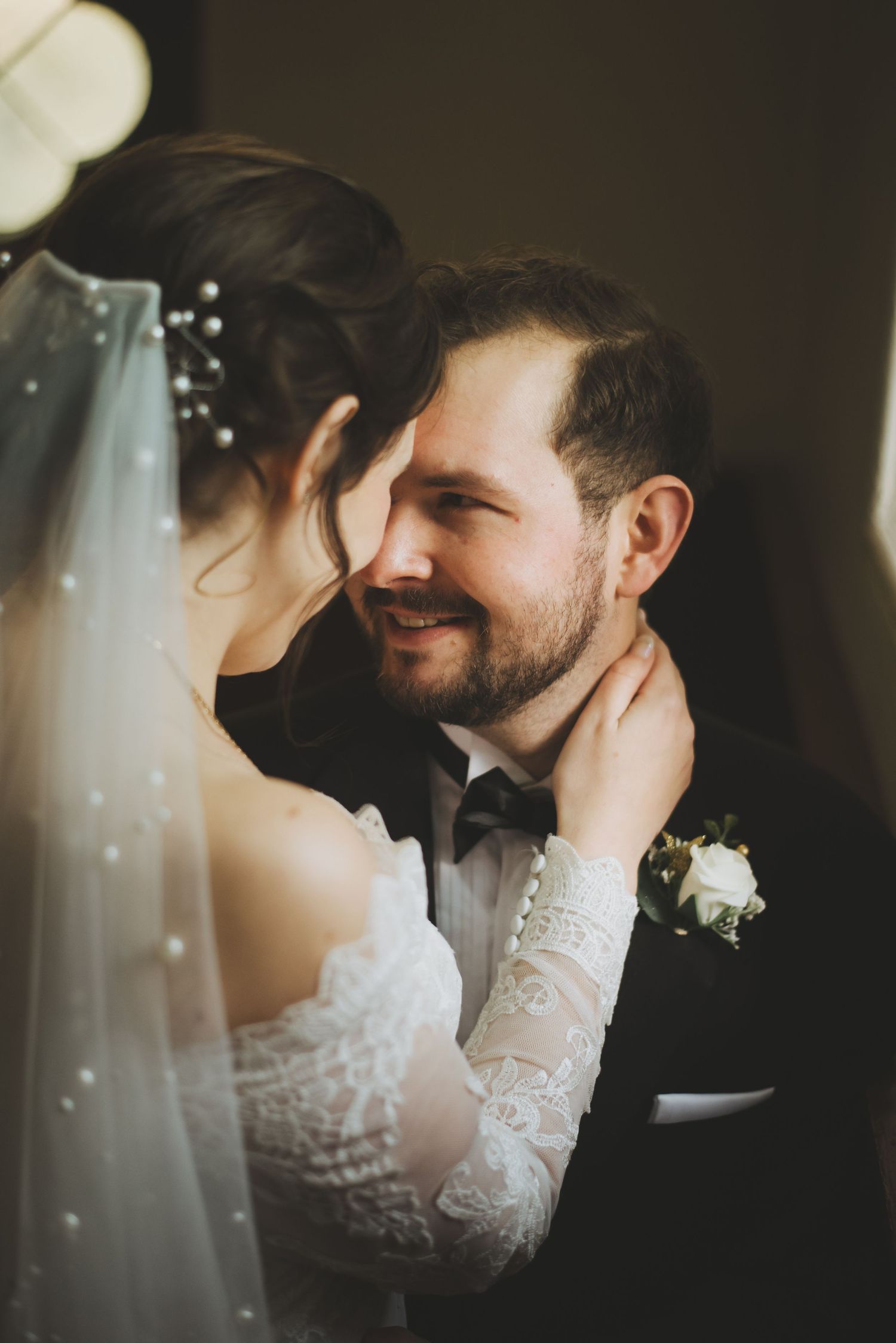 Intimate wedding moment captured as bride in lace sleeves and veil caresses groom wearing bowtie and boutonniere.