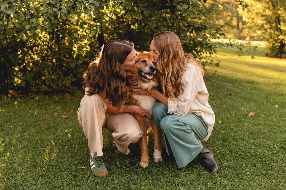 A golden retriever dog sits on grass between two people in a sun-dappled outdoor setting during golden hour.