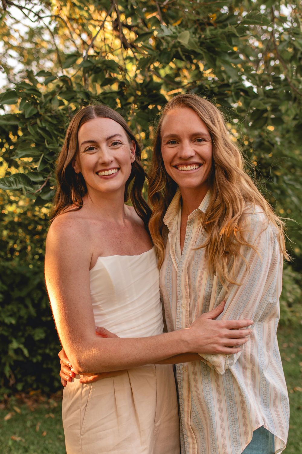 Two woman embracing and smiling together outdoors during golden hour with trees in background.