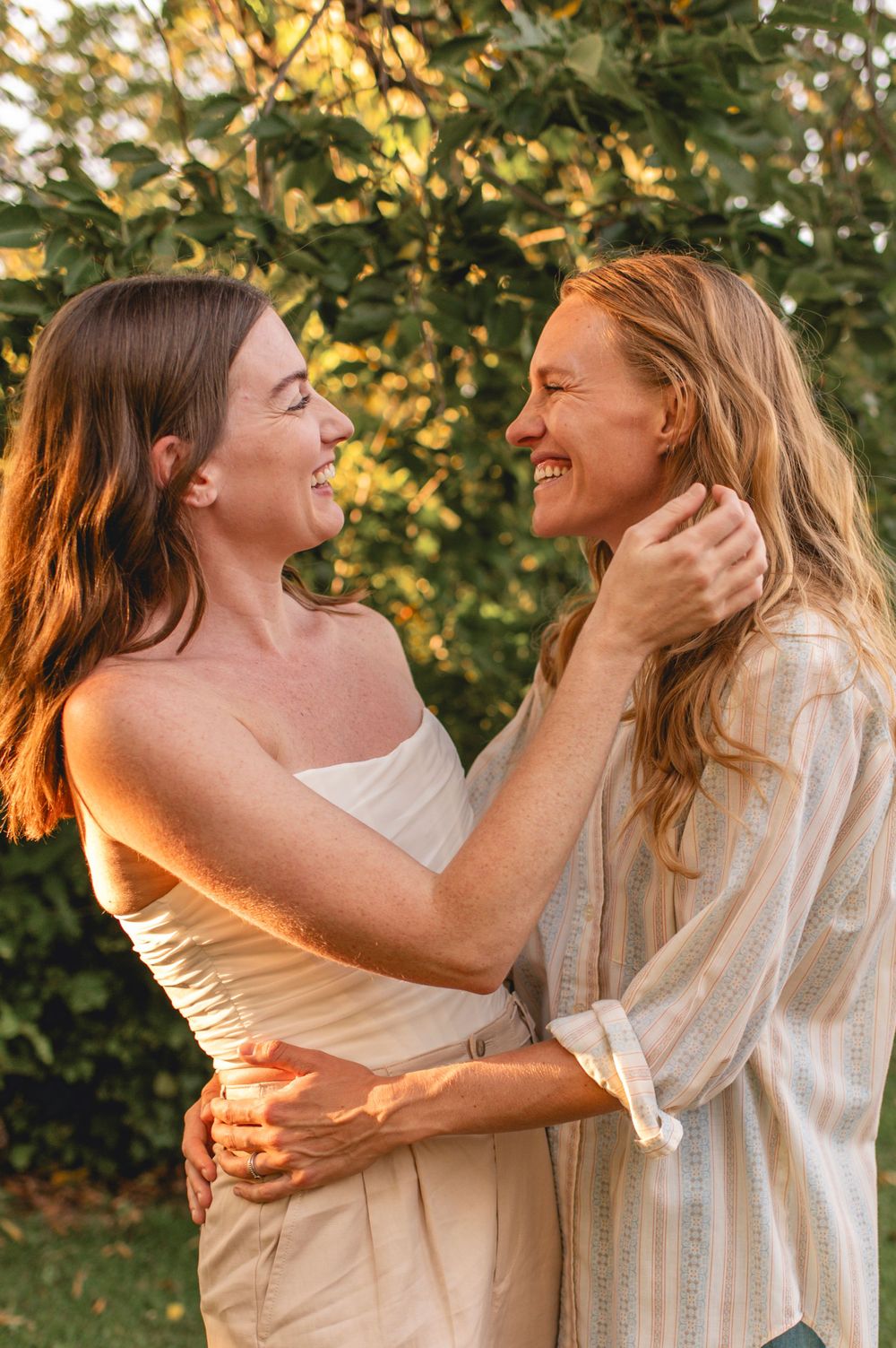 Two people share a joyful embrace while laughing together in golden sunlight against green foliage.