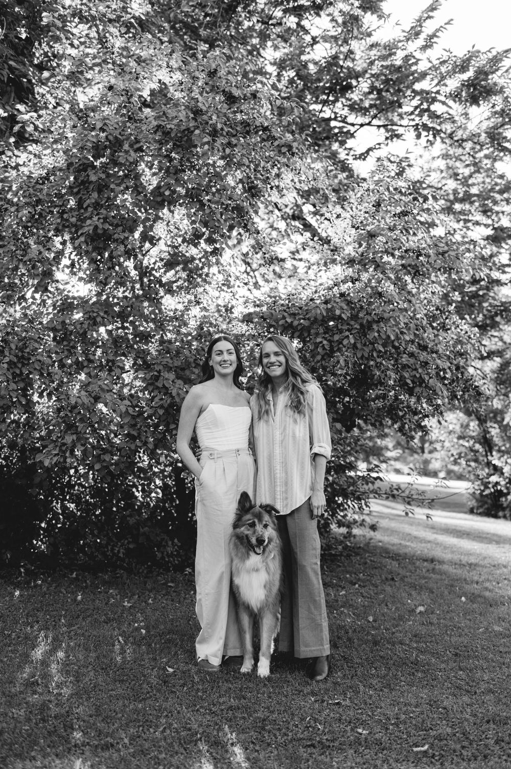 A black and white photograph of two people standing outdoors with a collie dog under a leafy tree canopy.