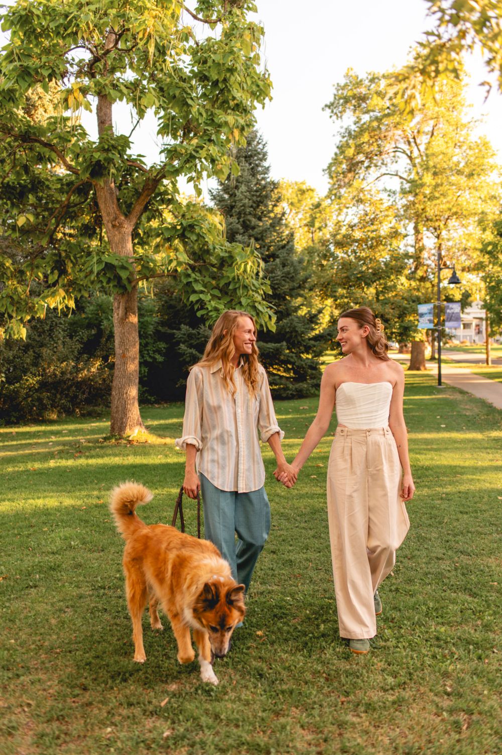 Two companions stroll through a sunny park with their fluffy brown dog, wearing casual and formal attire.