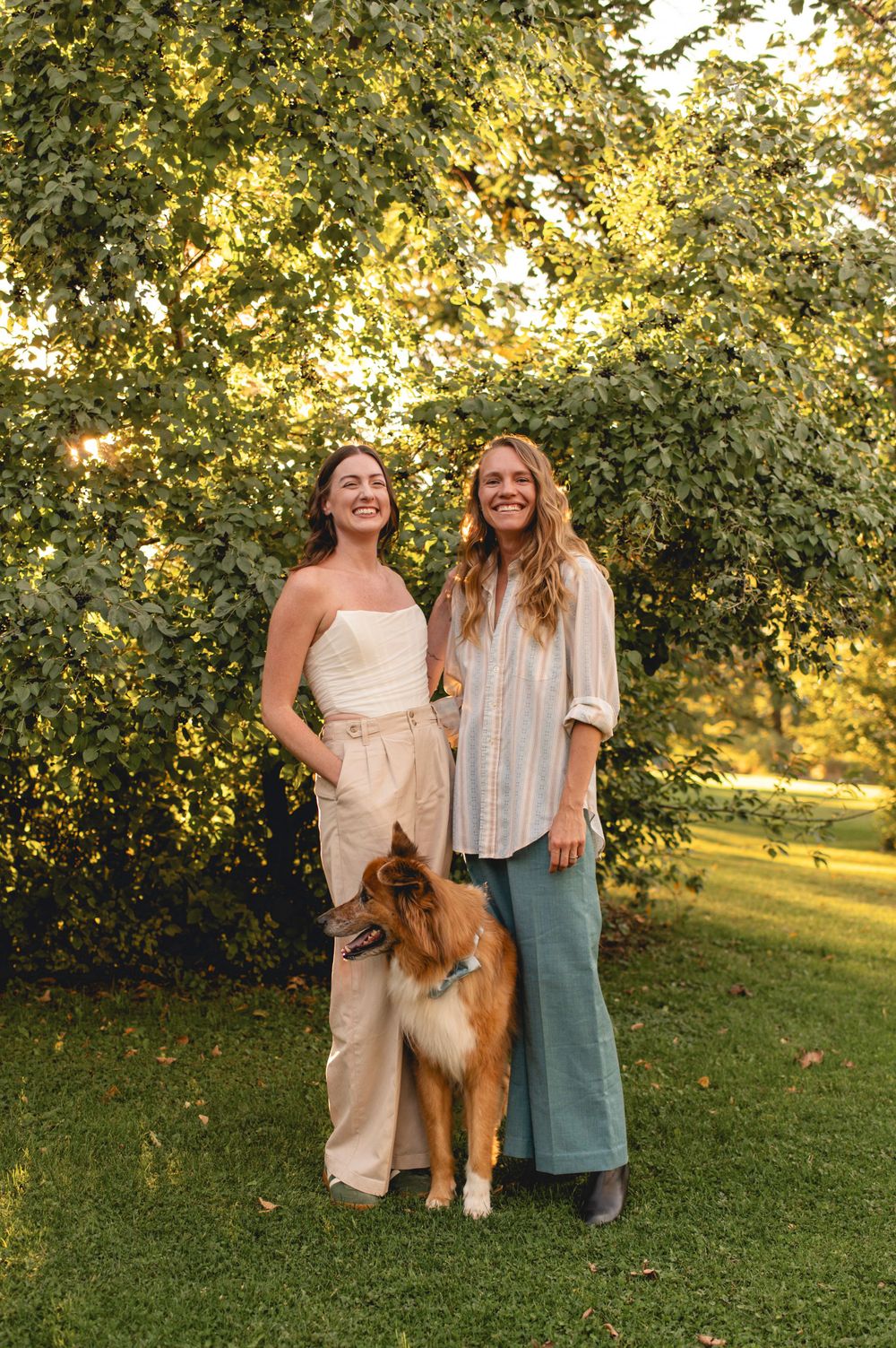 Two people stand together with a collie dog on green grass with sunlit trees in the background during golden hour.