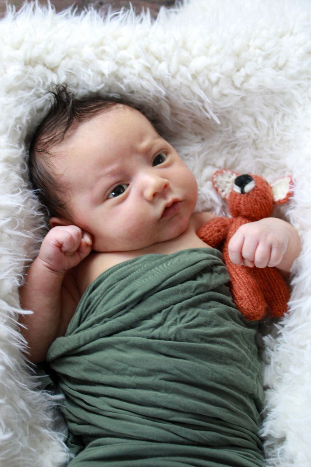 Baby in olive green wrap cuddling small orange fox toy on white fur.