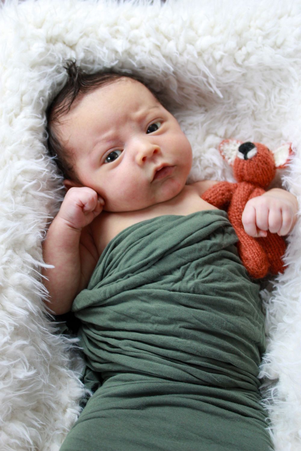 Baby wrapped in green fabric holding orange fox toy on white fur backdrop.