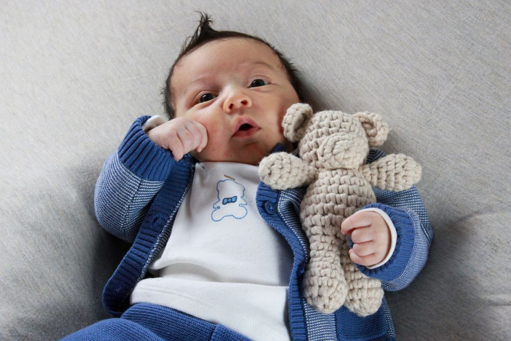 Baby in blue cardigan lies on grey surface holding a plush teddy bear toy.