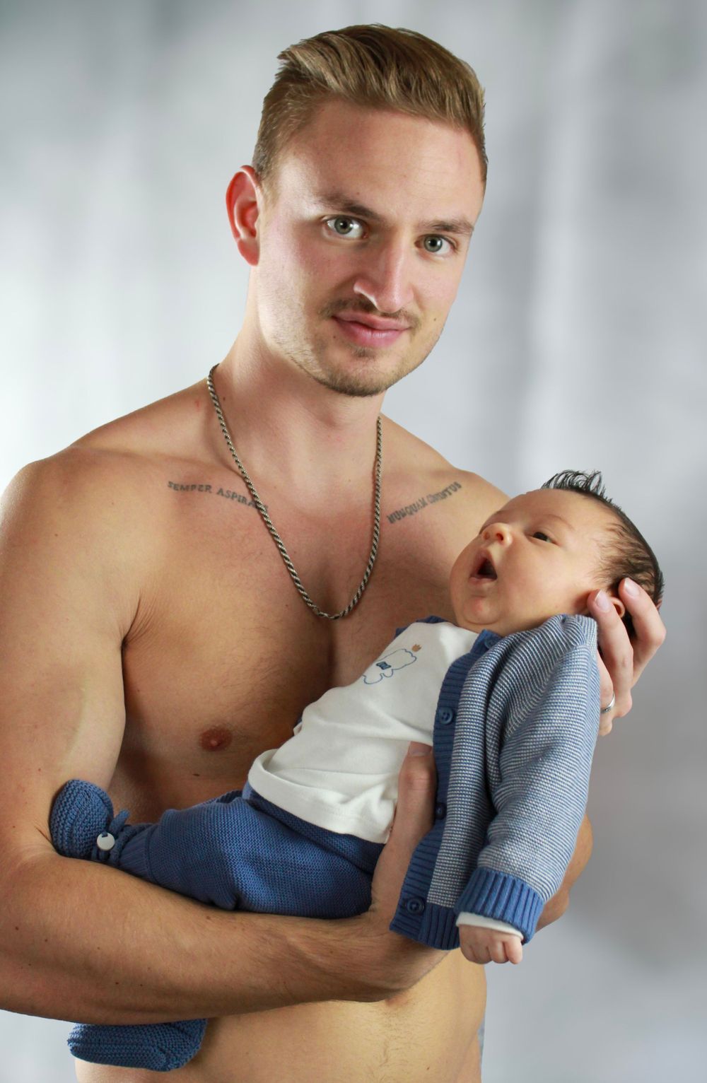 A shirtless parent tenderly holds an infant wearing a blue cardigan against a light background.