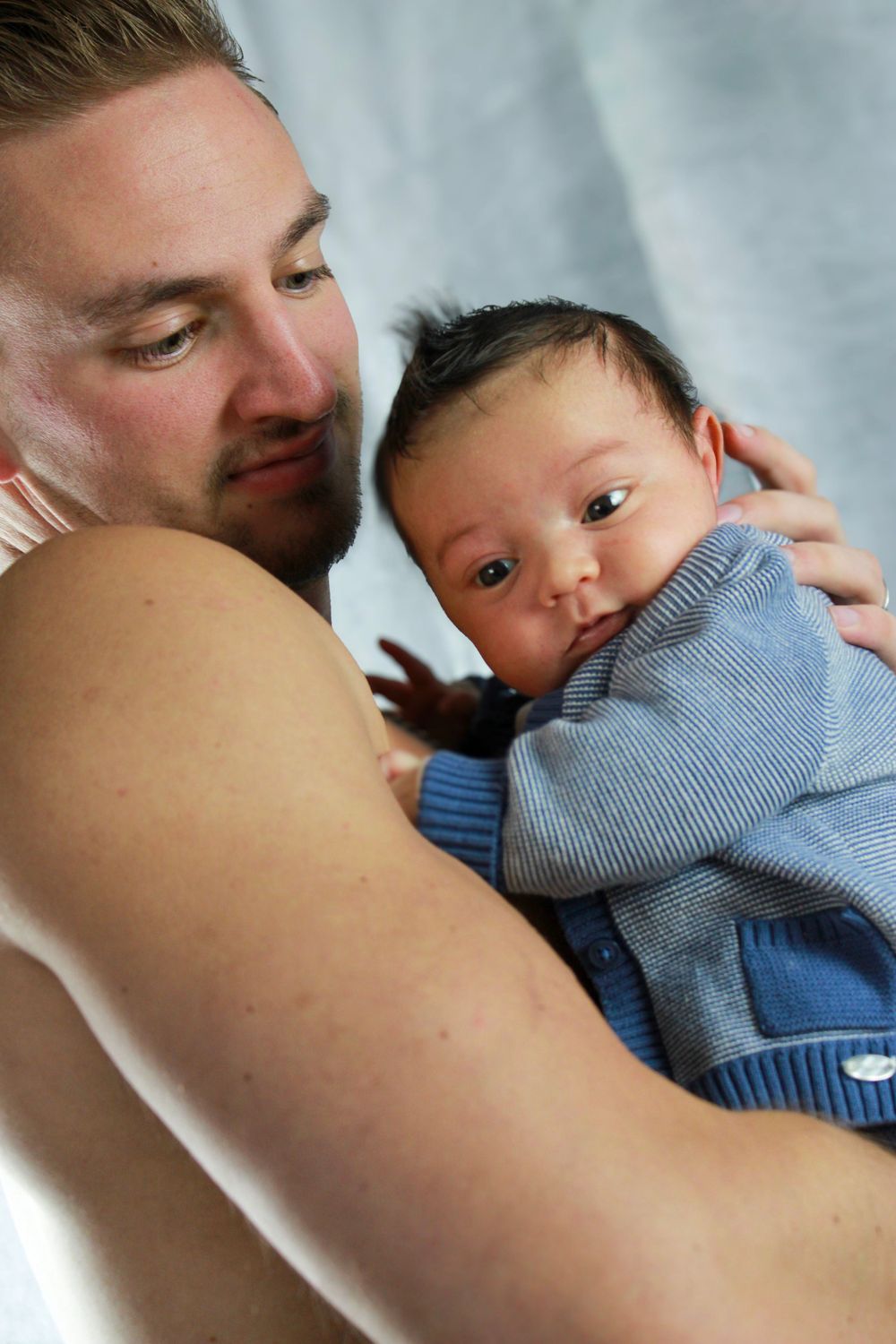 Parent lovingly embraces baby wearing blue sweater in intimate portrait against white backdrop.
