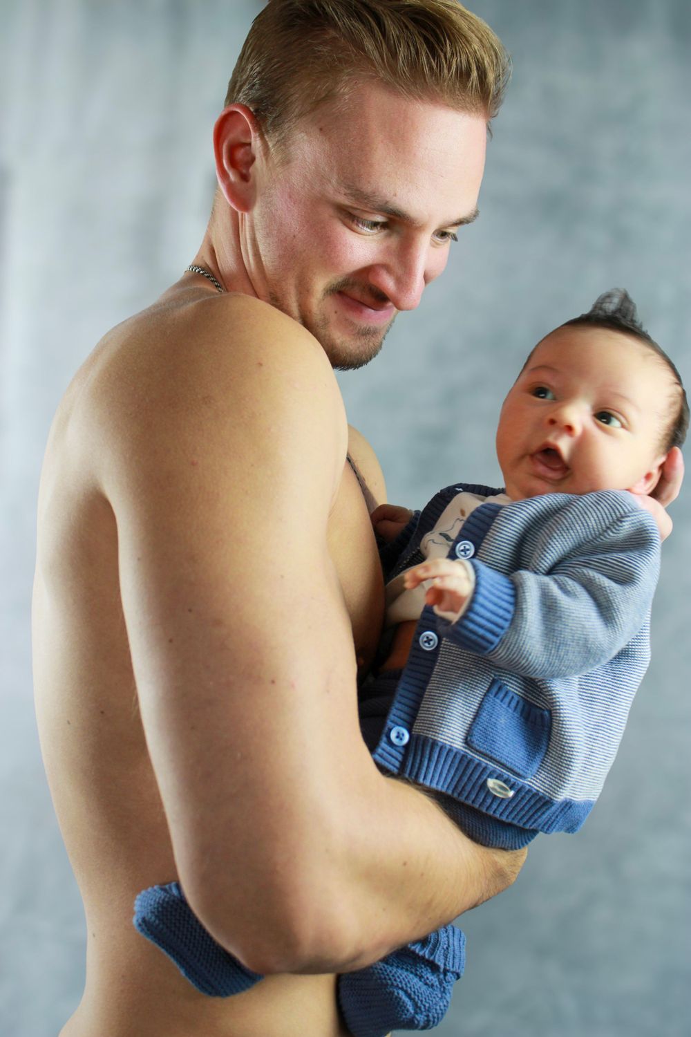Parent cradles infant wearing blue sweater in tender portrait against light background.