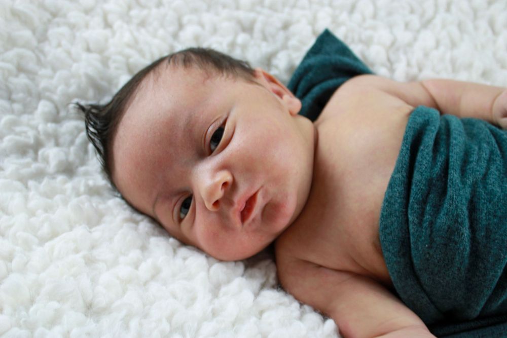 A newborn wrapped in teal fabric rests on a white fluffy blanket.