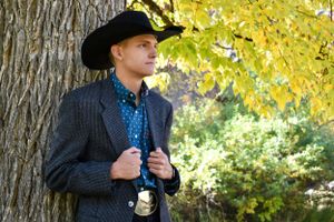 A man wearing a cowboy hat and blazer leaning against a tree and looking into the distance.