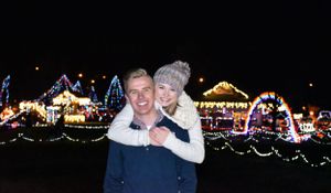 A couple posing in front of a park full of christmas lights.