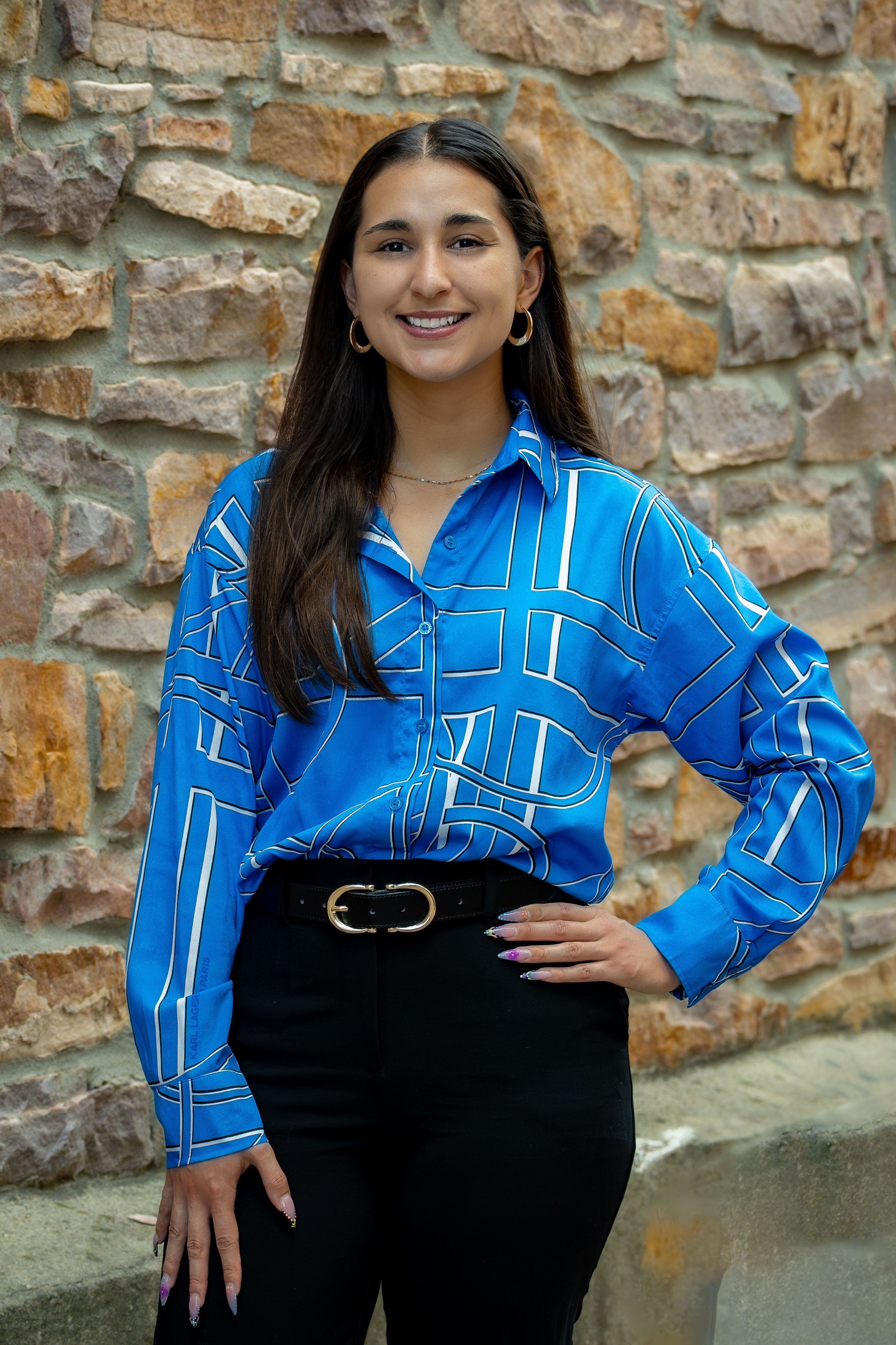 A woman wearing a bright blue top and black pants posing for a professional headshot in front of a brick wall.