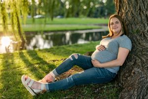 A pregnant woman sitting at the base of a willow tree with her hands on her stomach.