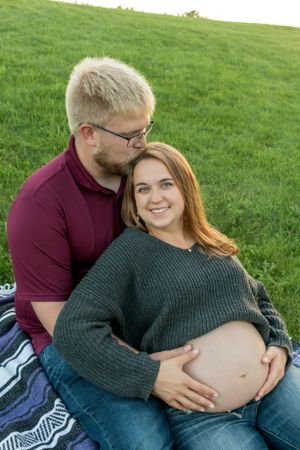 A couple sitting on a hillside with the woman's pregnant belly out.