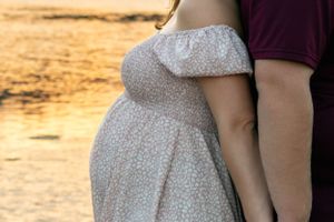 A couple standing back to back in front of a lake reflecting the sunset. The woman is pregnant.