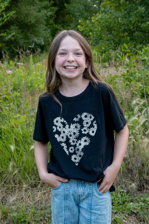 A young girl smiling at the camera wearing blue jeans and a black t-shirt with a heart made of flowers.