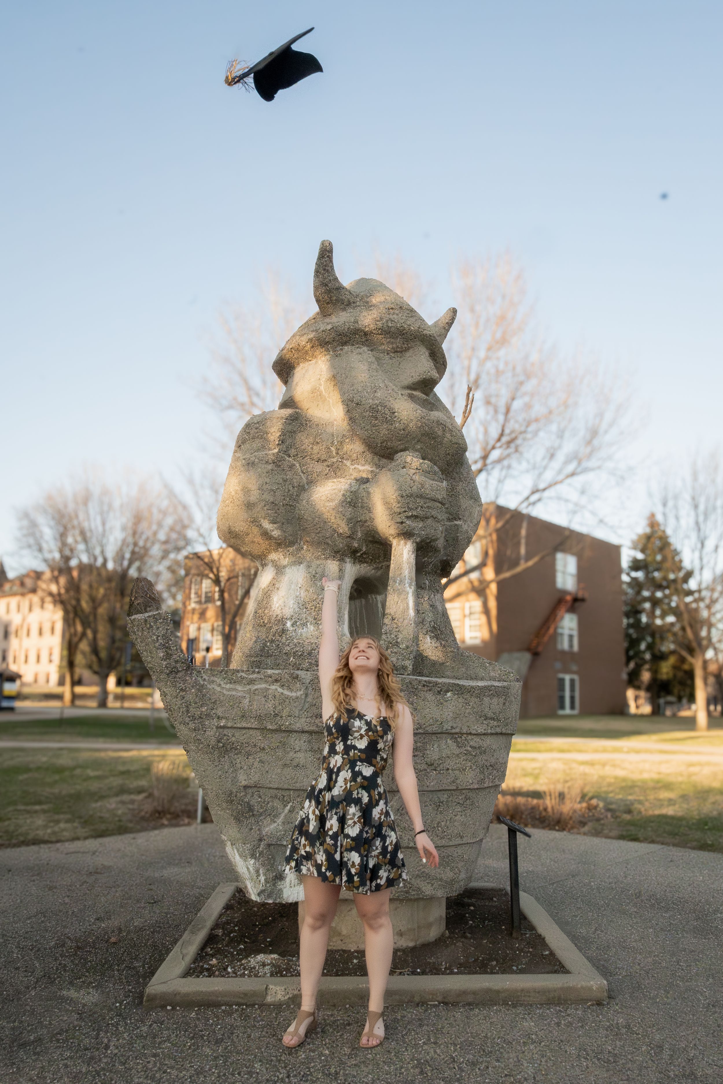 A young woman throwing a grad cap in the air in front of a statue of her mascot.
