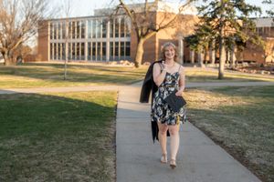 A young blonde woman in a dress carrying a graduation cap and gown walking outdoor on a college campus.