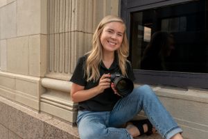 A woman sitting in a windowsill holding a camera and smiling.