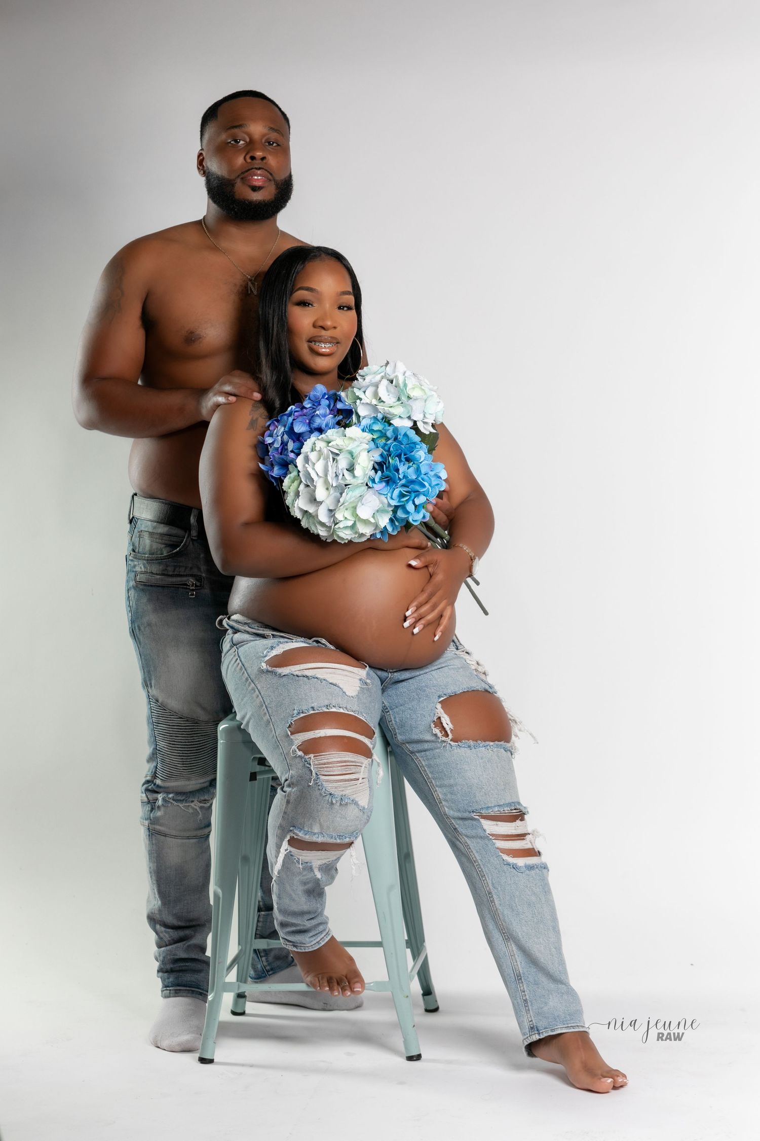 Artistic maternity photoshoot featuring a couple in distressed jeans with blue hydrangea flowers.