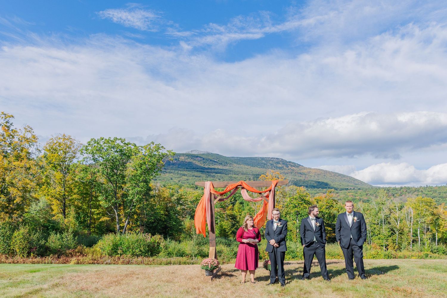 Wedding party stands under a rustic wooden arch with coral draping and mountain views.