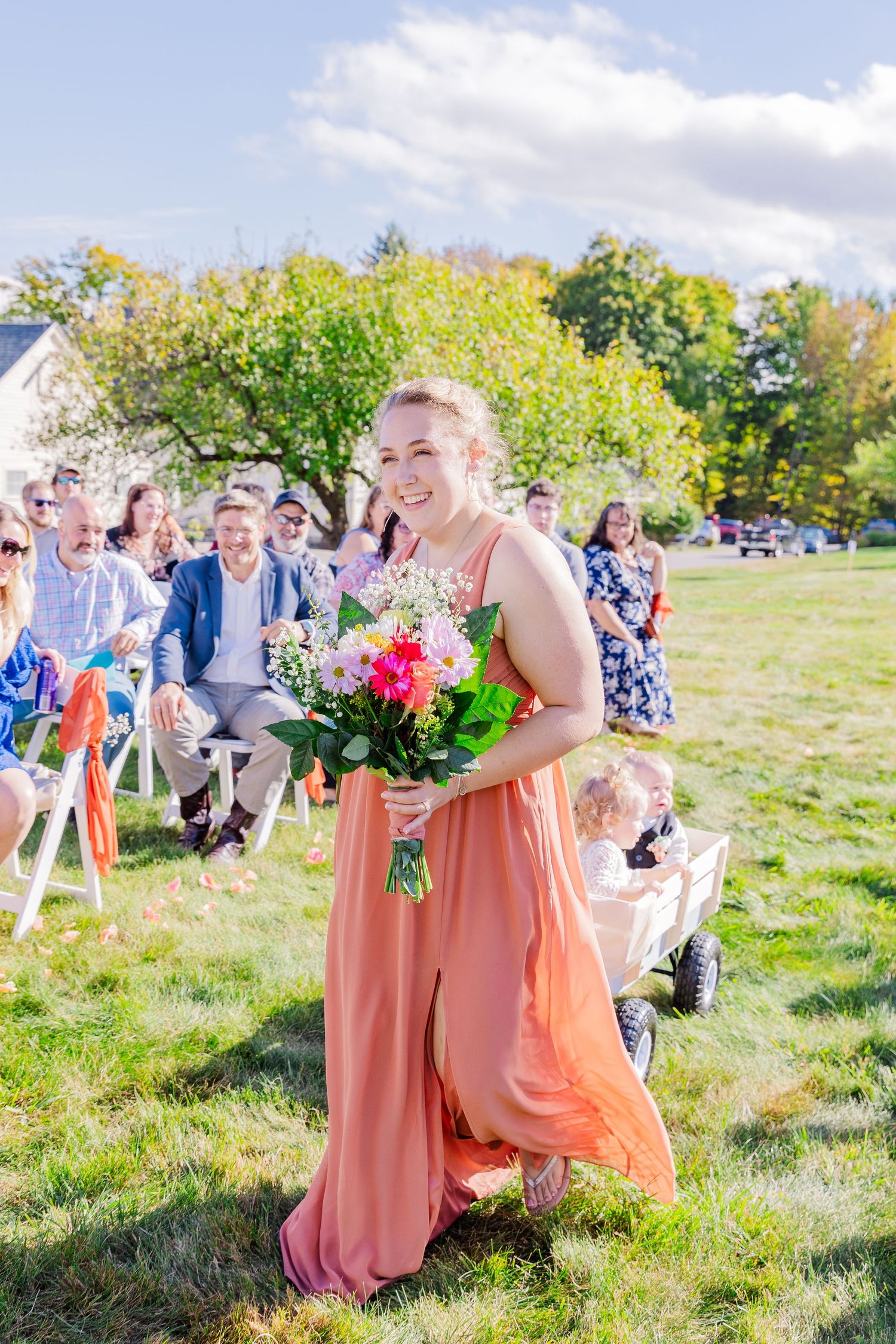 Attendant in a coral dress walks down the aisle holding vibrant flowers during an outdoor ceremony.