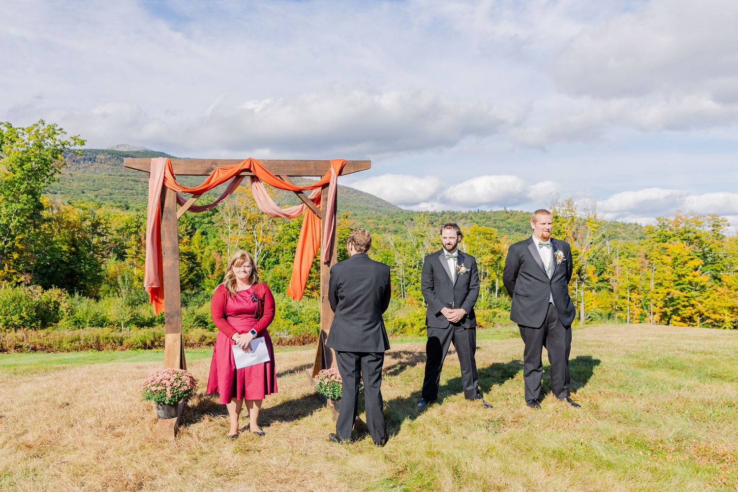 Wedding ceremony setup with wooden arch draped in coral fabric against mountain backdrop.